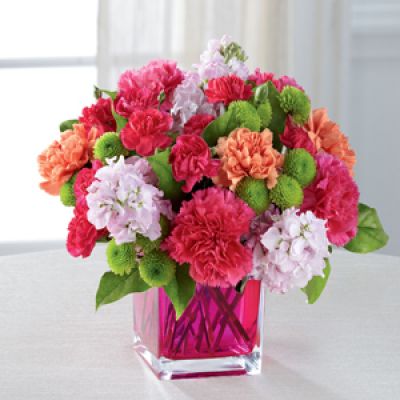 Dazzling and delightful, this bold and colorful flower bouquet is set to impress with it's high impact look and graceful styling. Hot pink carnations, orange carnations, pale pink gilly flower, hot pink mini carnations, green button poms, and lush greens are beautifully arranged in a raspberry pink glass cubed vase to create a rush of fun and warm wishes to send straight to your recipient's door.